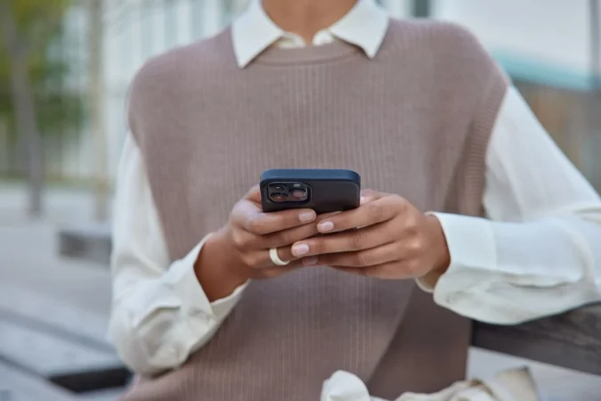 woman elegant clothes holds mobile phone surfs wireless internet makes shopping online sits bench outside connected wireless internet communicates distantly Merca2.es