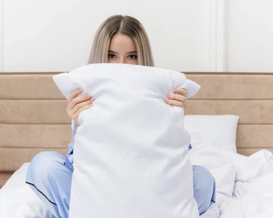 young beautiful woman blue pajamas sitting bed with pillow hiding face peeking bedroom interior Merca2.es