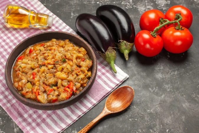 vista superior de la ensalada de berenjena asada en un tazon de madera cuchara tomates berenjenas botella de aceite en superficie oscura Merca2.es