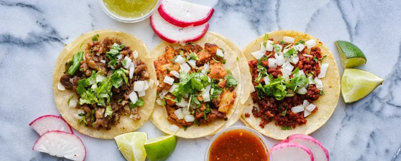 fila de tacos callejeros mexicanos surtidos con guarniciones en una amplia composicion de banner Merca2.es