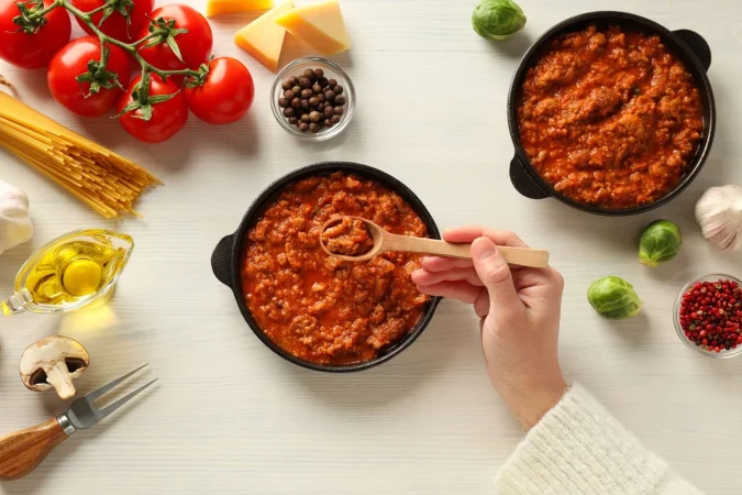 sartenes con salsa bolognese cuchara de madera en la mano e ingredientes en fondo de madera blanca vista superior Merca2.es