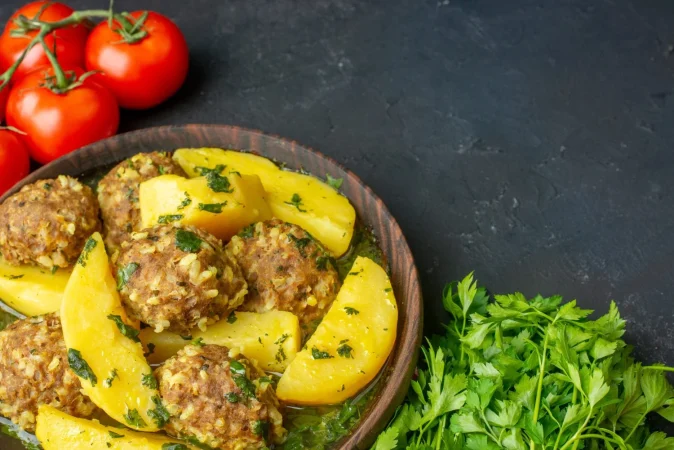 vista de cerca de una cena sabrosa con albondigas papas verdes en un tazon marron sobre tomates de toalla pelados rojos sobre fondo negro con espacio libre Merca2.es