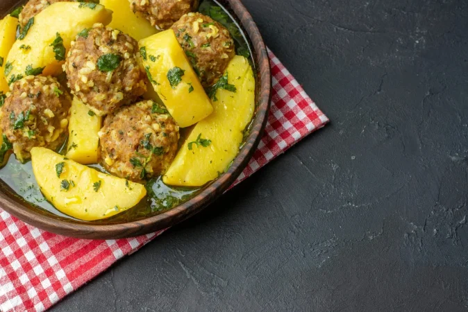 vista superior de una cena sabrosa con albondigas papas verdes en un tazon marron sobre una toalla roja pelada sobre fondo negro con espacio libre Merca2.es